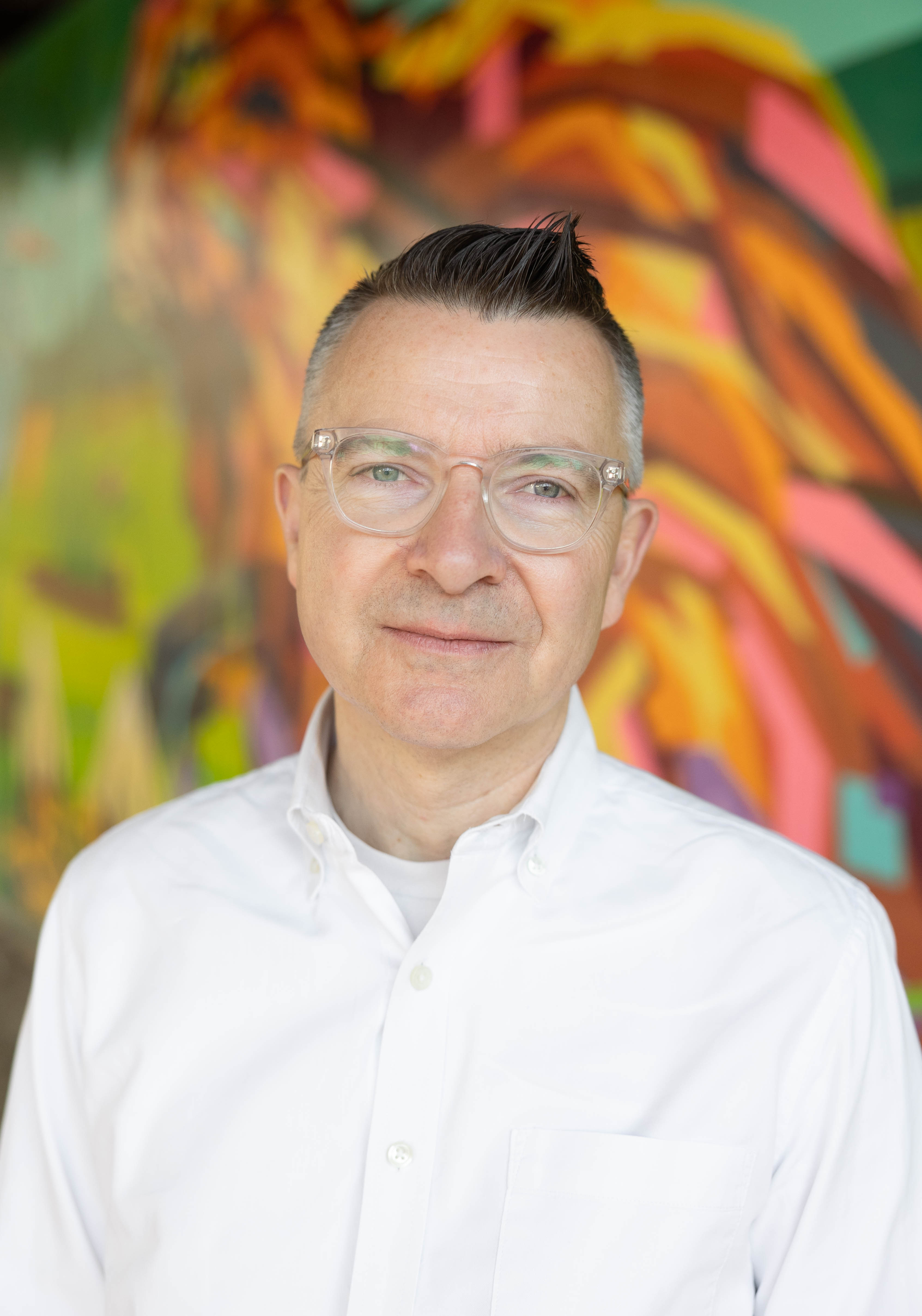 Greg Peckham, CEO of Conservancy for Cuyahoga Valley National Park, wearing a white button-down shirt and clear-framed glasses, stands in front of a colorful abstract mural, smiling at the camera.