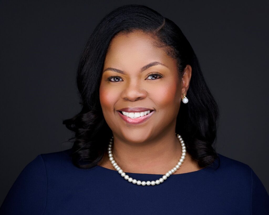 Professional headshot of Tracy Carter, president and CEO of Trailhead Community Health Foundation, smiling against a dark background. She wears a navy blue dress, pearl necklace and pearl earrings.