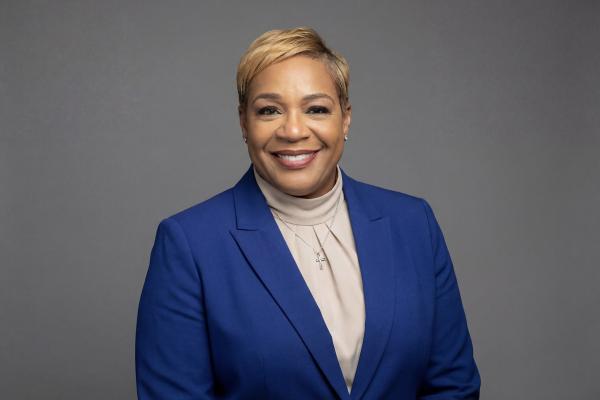 Professional headshot of a woman in a blue blazer smiling against a gray background.
