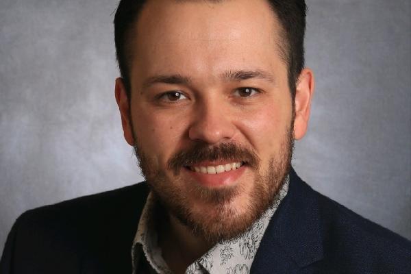Caucasian man with short dark hair and beard wearing suit and tie smiling into camera.