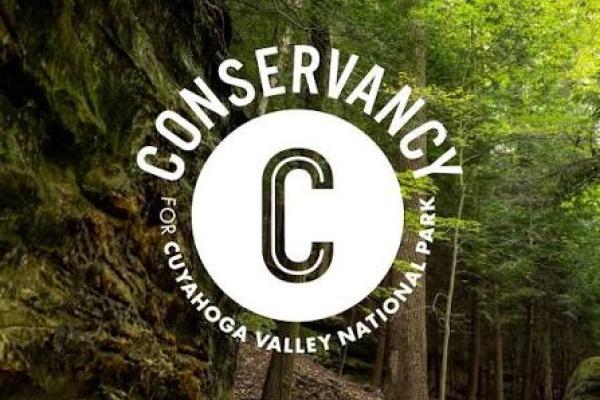 Conservancy for Cuyahoga Valley National Park logo over a wooded park trail bordered by moss-covered rock cliffs and tall trees.