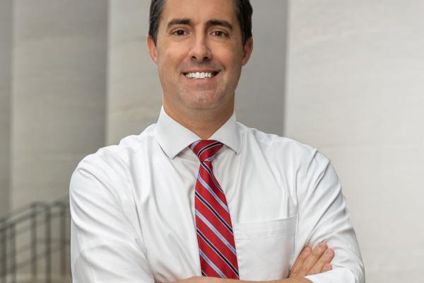 Professional headshot of a man in a white shirt and red striped tie, smiling with arms crossed.