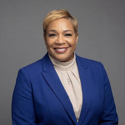 Professional headshot of a woman in a blue blazer smiling against a gray background.