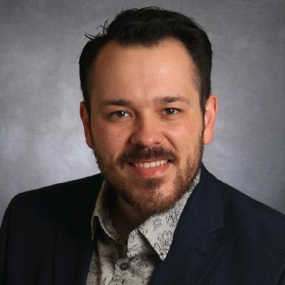Caucasian man with short dark hair and beard wearing suit and tie smiling into camera.
