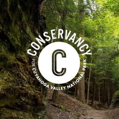 Conservancy for Cuyahoga Valley National Park logo over a wooded park trail bordered by moss-covered rock cliffs and tall trees.