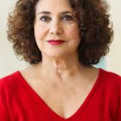 Middle-aged white woman with dark brown curly hair wearing a red dress and smiling into the camera.