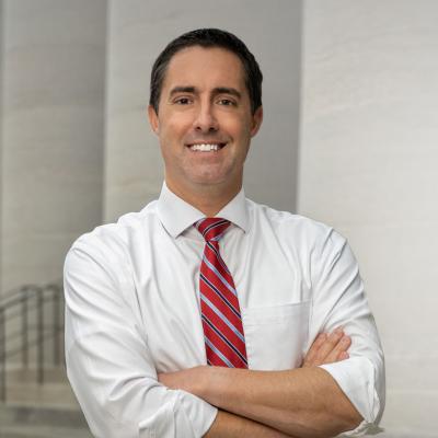 Professional headshot of a man in a white shirt and red striped tie, smiling with arms crossed.